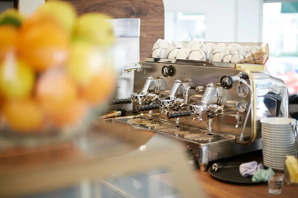 Die Faema-Espressomaschine auf dem Tresen im Café Délice, darauf gestapelte Tassen, im Vordergrund eine Schale mit Zitrusfrüchten